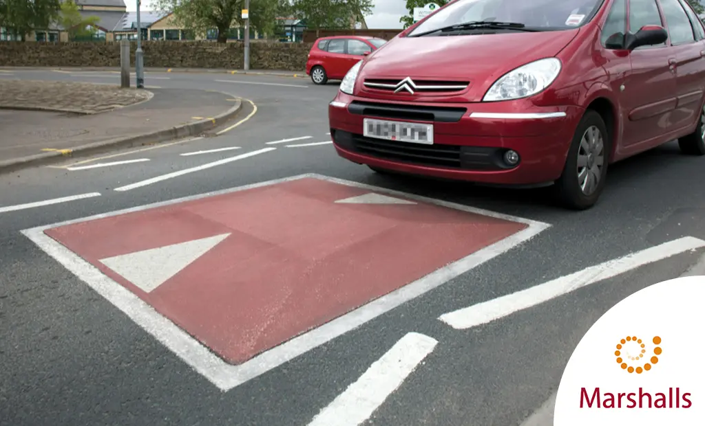 A red car is about to pass over a red and white concrete road cushion made by Marshalls.